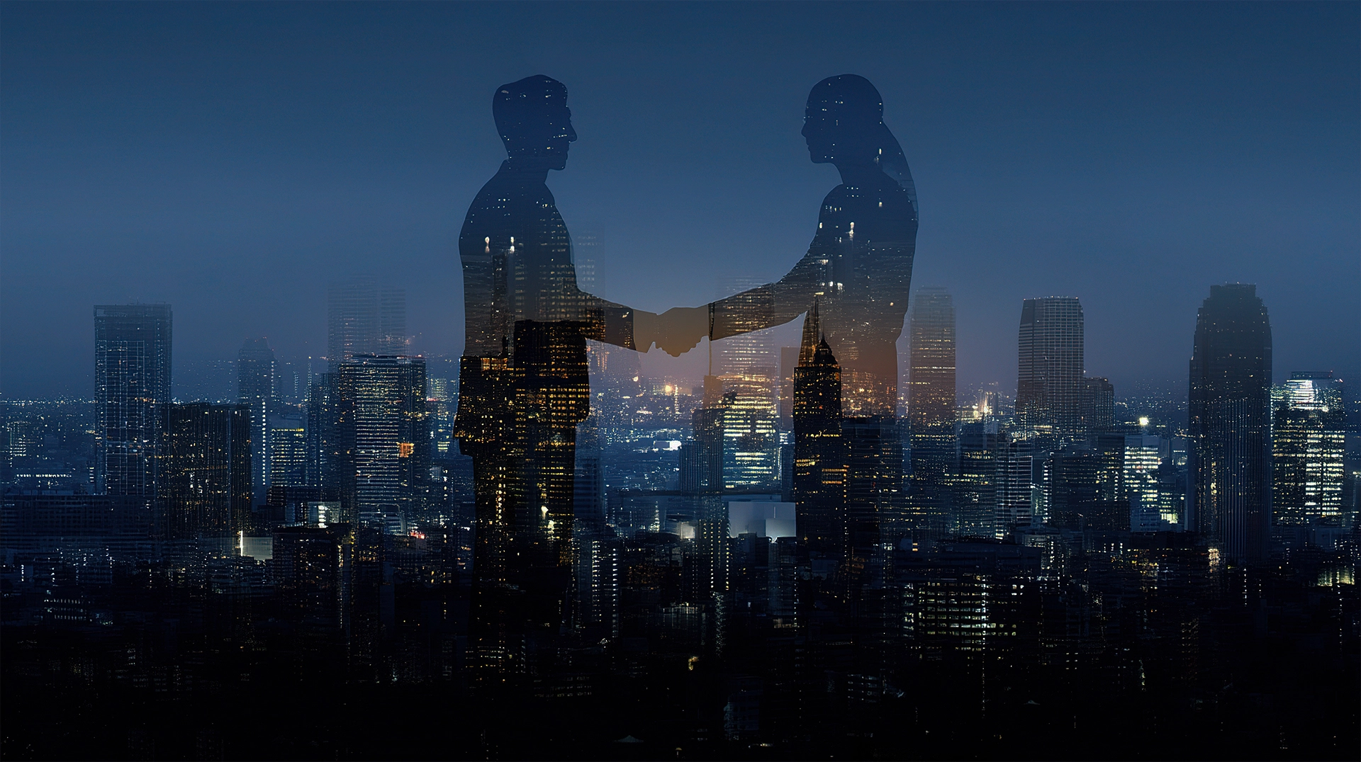 Silhouettes of two business professionals shaking hands over a city skyline at dusk with building lights illuminated.